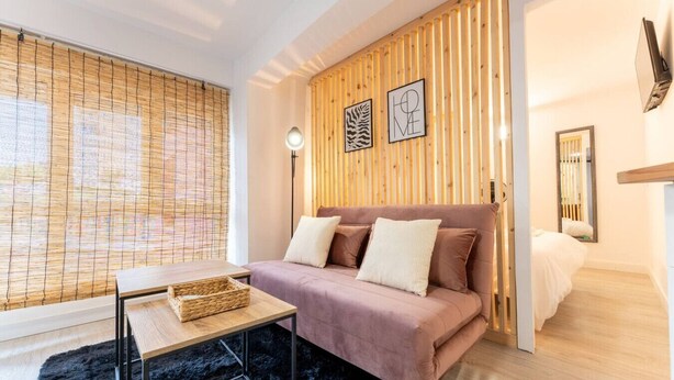 Ein modernes Schlafzimmer mit einer Wand aus Holzlatten, einem Bett mit rosa Bettdecke, einem kleinen Tisch mit Korb und einem schwarzen, flauschigen Teppich.