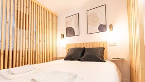 Ein ordentlich bezogenes Bett mit weißen Leinen und zwei dunklen Kissen, eine Wand aus Holzlatten sowie zwei abstrakte Kunstwerke an der Wand.