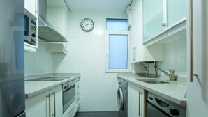A compact kitchen with white cabinets, a built-in oven, a microwave, a sink, and a clock on the wall. - Fuentes de encanto en Santander 2 hab. (Santander)