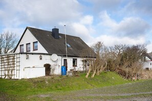 Une maison blanche avec un toit en pente, une antenne parabolique et une poubelle bleue dans la cour.