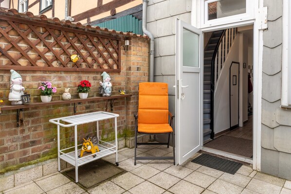 Rincón de una habitación con pared de ladrillo, estante metálico blanco con objetos decorativos, una silla naranja y una puerta blanca que da a una escalera.