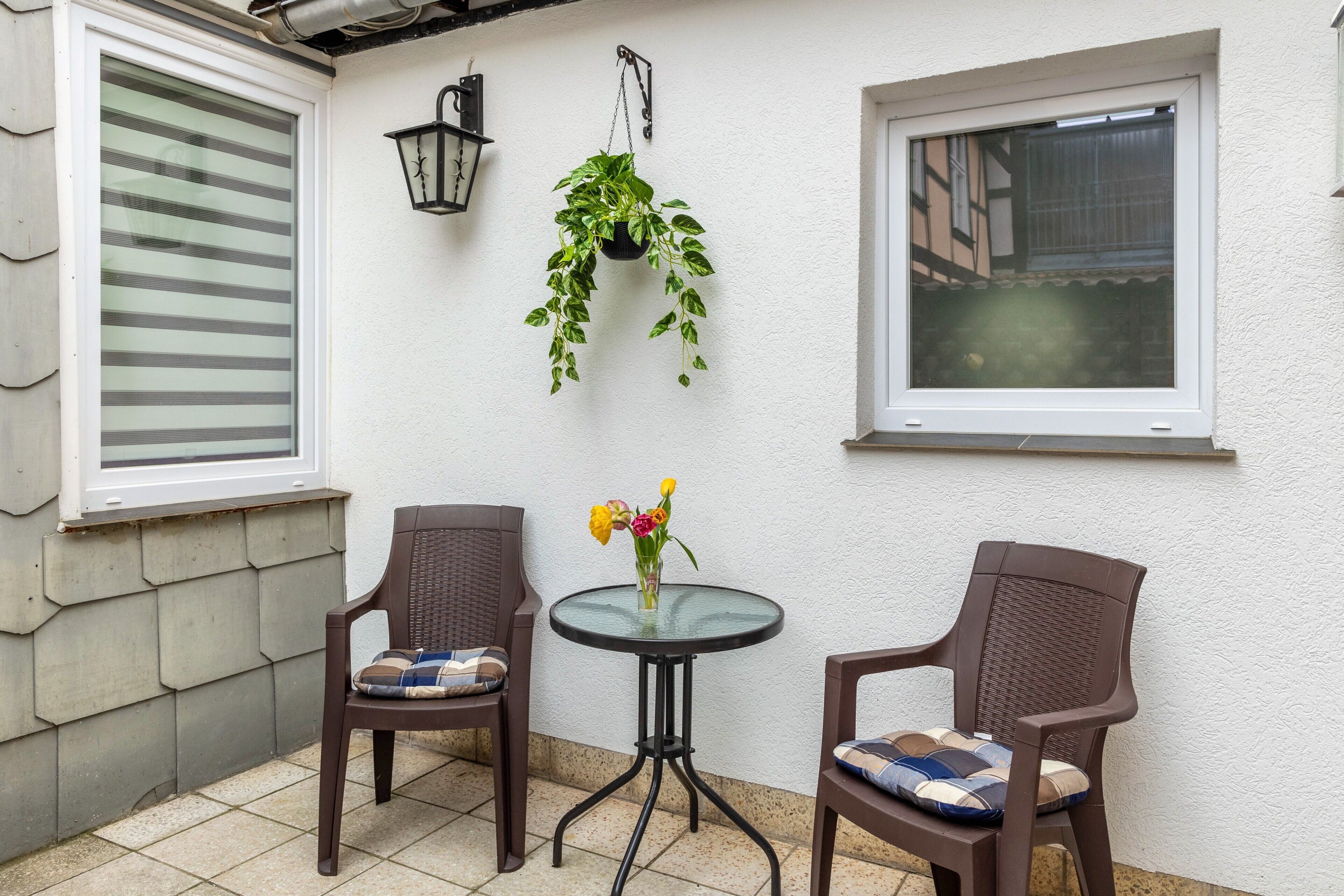 A small patio area with two wicker chairs, a glass table, a hanging plant, and a window with blinds.