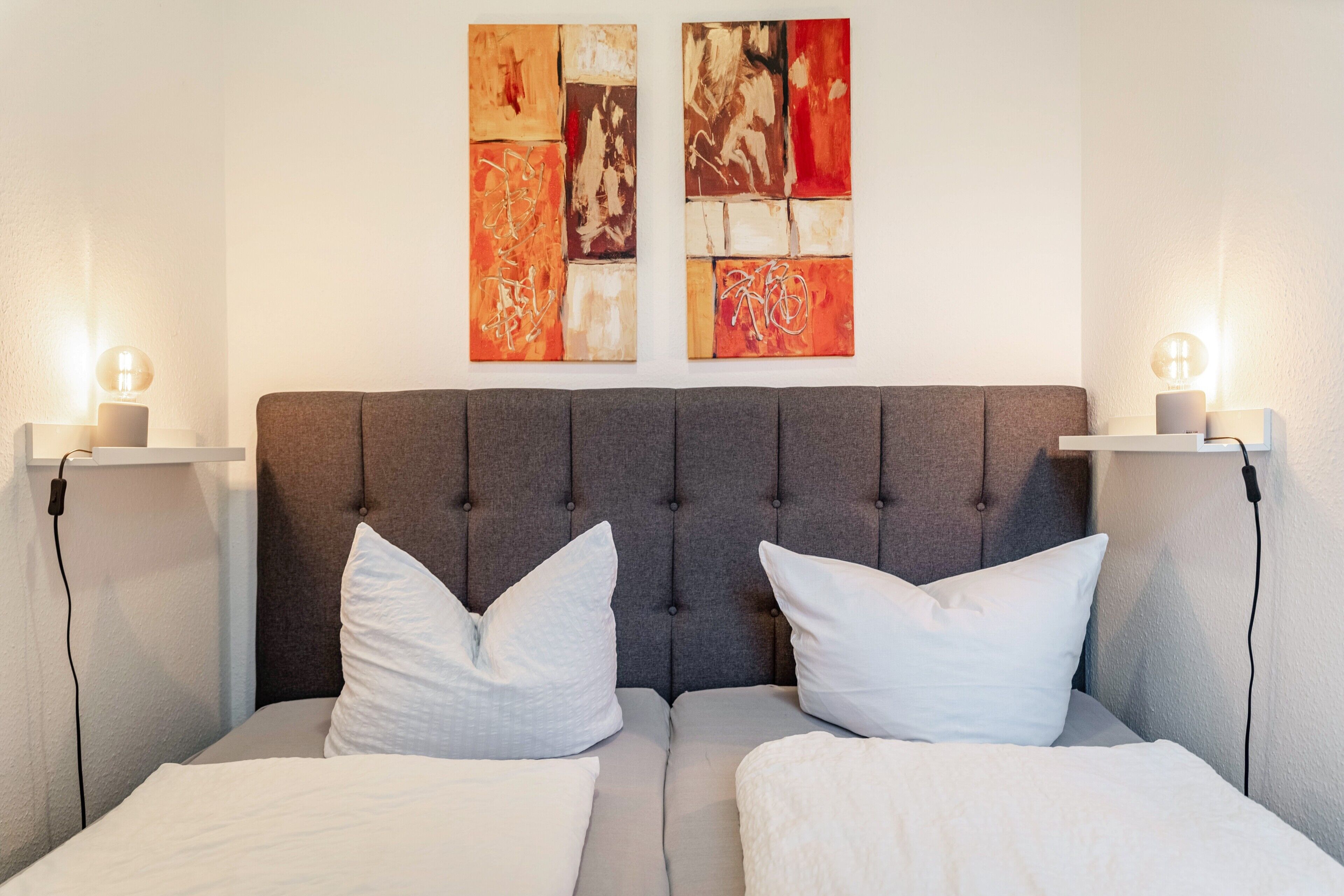 Un lit avec une tête de lit rembourrée de couleur grise, deux oreillers blancs et deux tables de chevet avec des lampes.