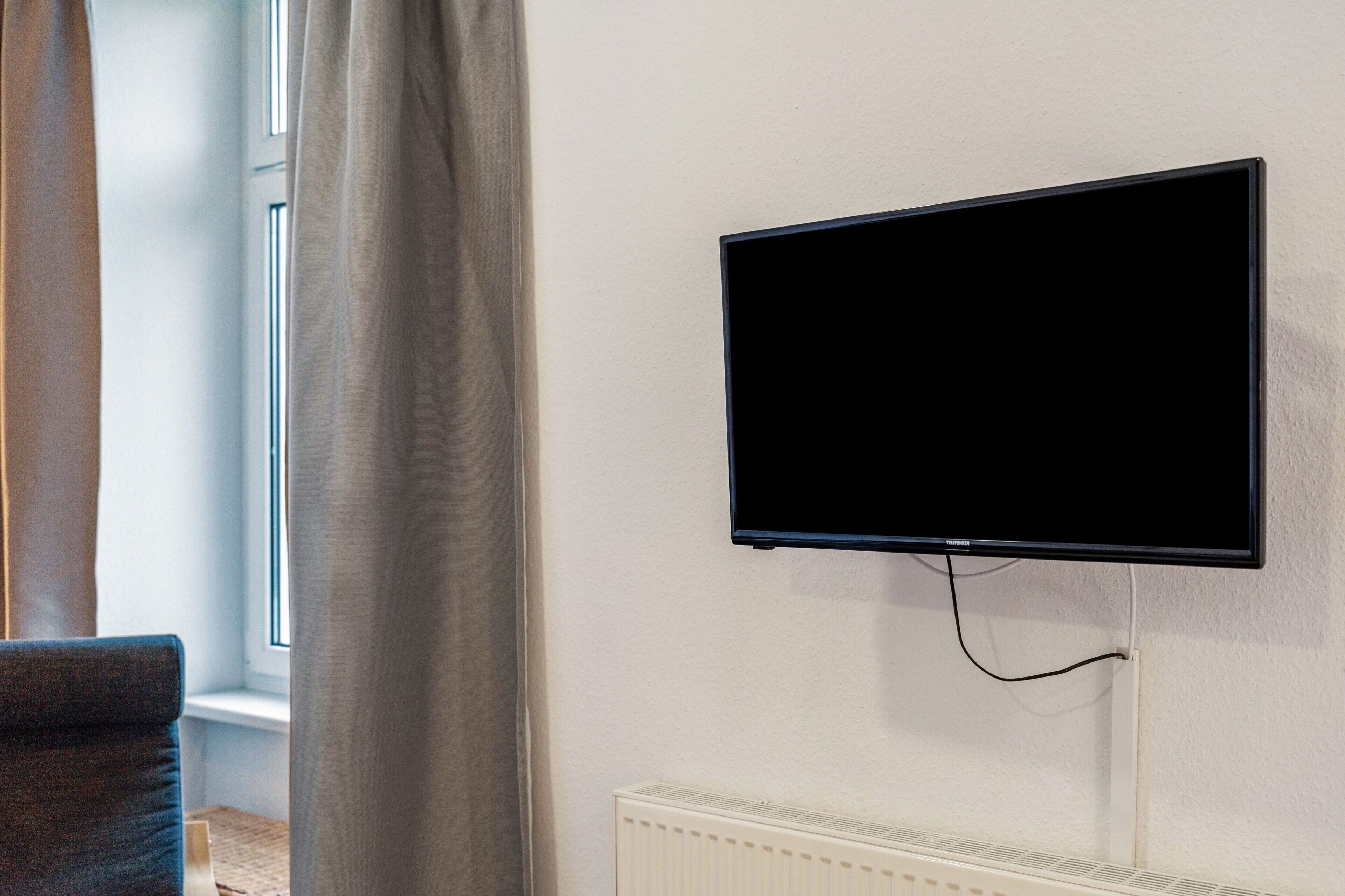 A room with a mounted TV on the wall, a window with curtains, and a radiator below.