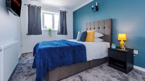 Une chambre à coucher avec un lit, un téléviseur fixé au mur, une table de chevet avec une lampe et une fenêtre avec des rideaux.