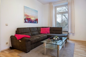 A modern living room with a sectional sofa, a glass coffee table, a painting on the wall, and a window with curtains. - Apartment 'Parkblick' with Shared Garden and Wi-Fi (Groß Plasten)