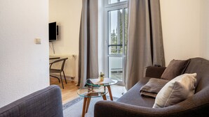 A living room with a sofa, a glass table, a radiator, a TV, and a window with curtains.