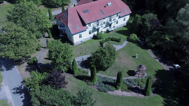 An aerial view of a large, two-story building with a red roof, surrounded by lush greenery and a well-maintained garden.