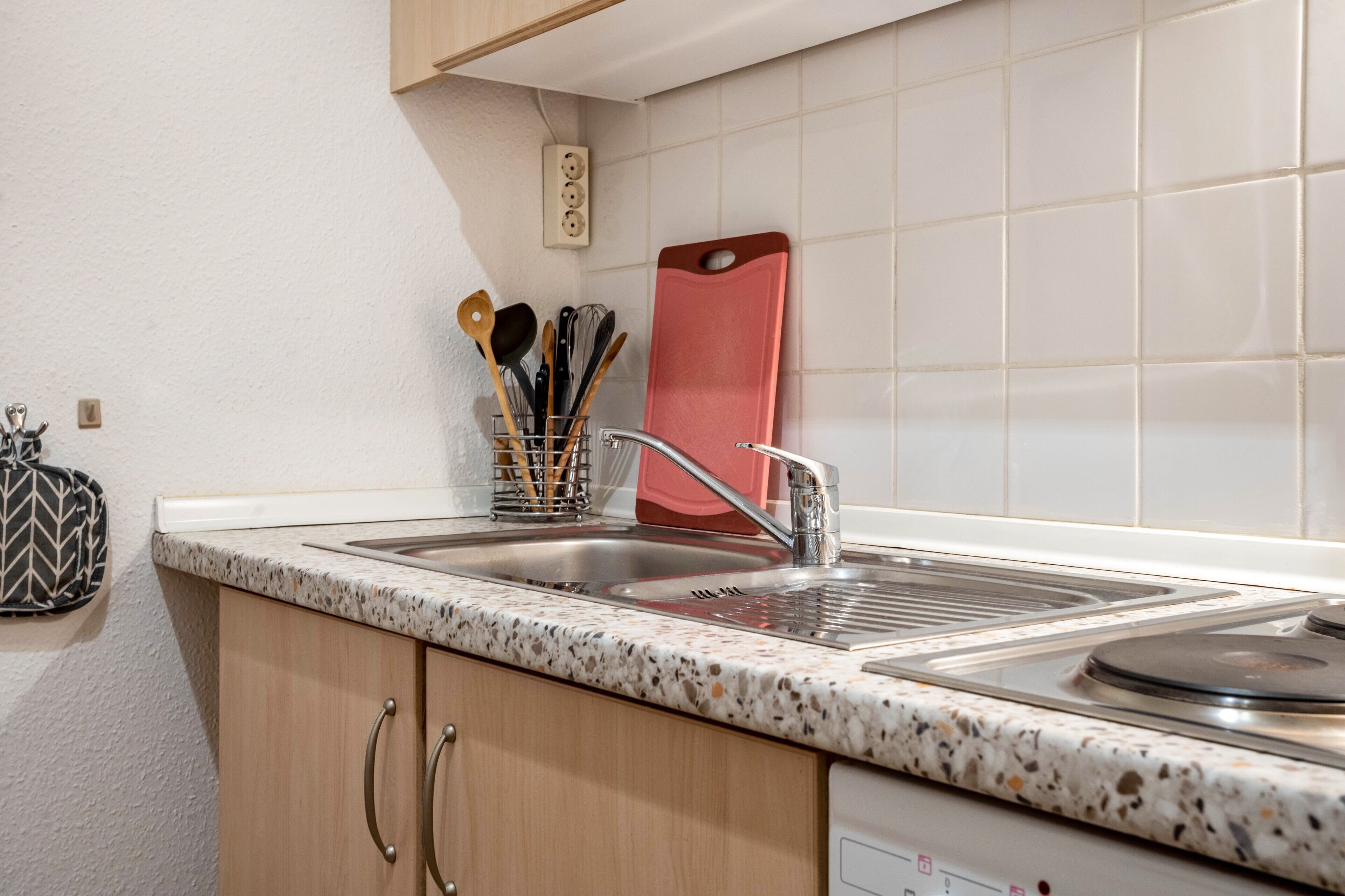 Een keuken met een dubbele spoelbak, houten kastjes, een rekje voor kruiden en een rood snijplank.