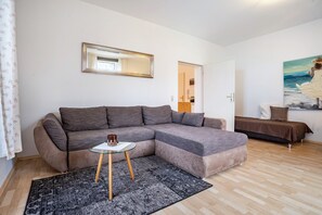 Ein modernes Wohnzimmer mit einem Ecksofa, einem kleinen Glastisch, einem gemusterten Teppich, einem Gemälde und einer Tür zu einem weiteren Raum.