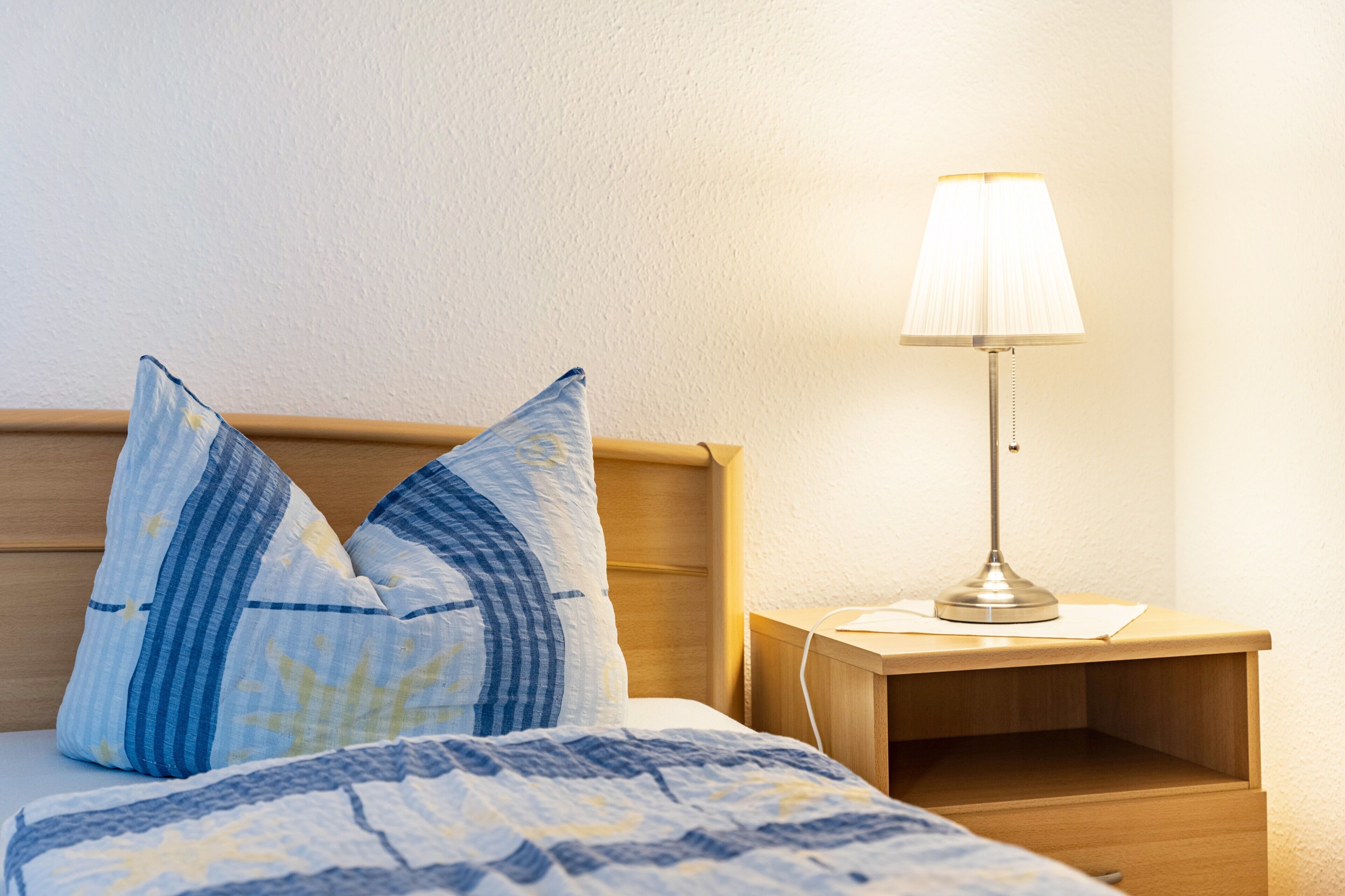 Un lit bien fait, avec une couvre-lit à motifs et deux oreillers, une table de chevet avec une lampe et une petite table en bois.