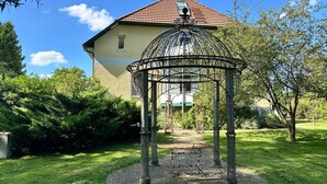En lusthus med ett tak och pelare av metall, omgivet av en rund stenlagd gång, framför ett hus med ett rött tegeltak.