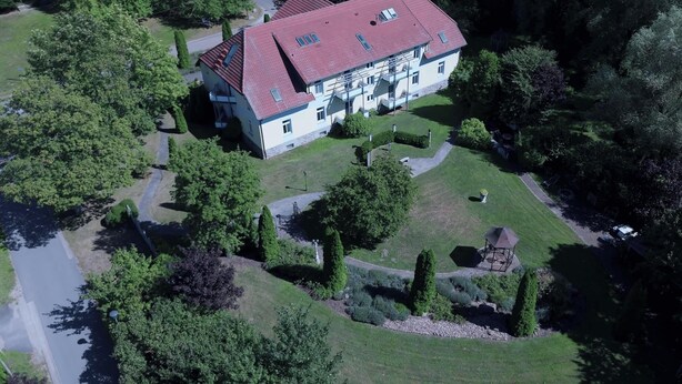 En vy från ovan av en stor byggnad i två våningar med ett rött tak, omgiven av frodig grönska och en välskött trädgård.