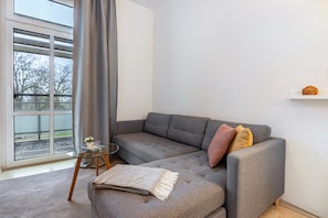 Ein modernes Wohnzimmer mit einem grauen Ecksofa, einem Glastisch, einem kleinen Beistelltisch, einer grauen Decke und Blick auf Bäume durch ein großes Fenster.
