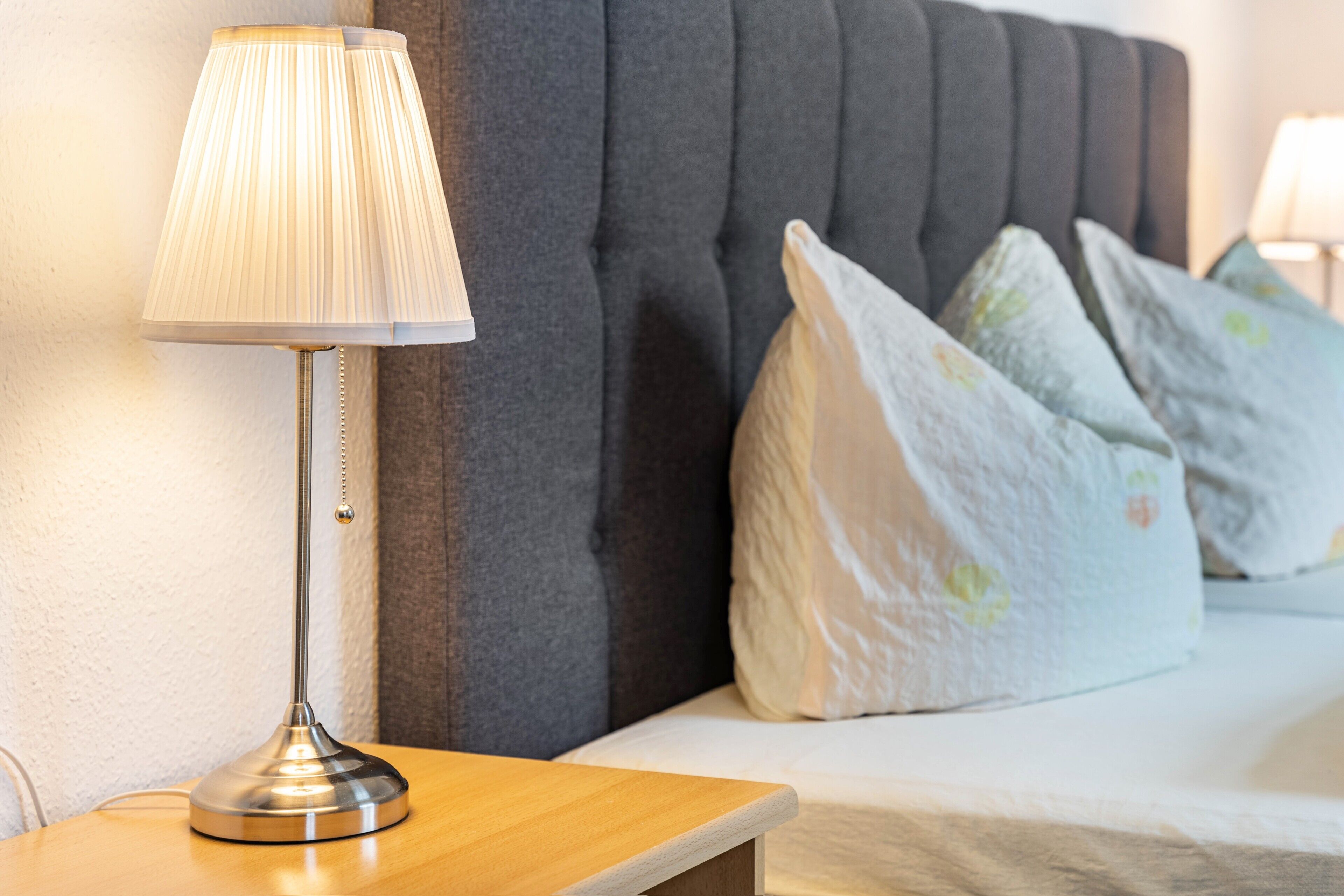 A bed with a grey upholstered headboard, a bedside table with a lamp, and patterned pillows.