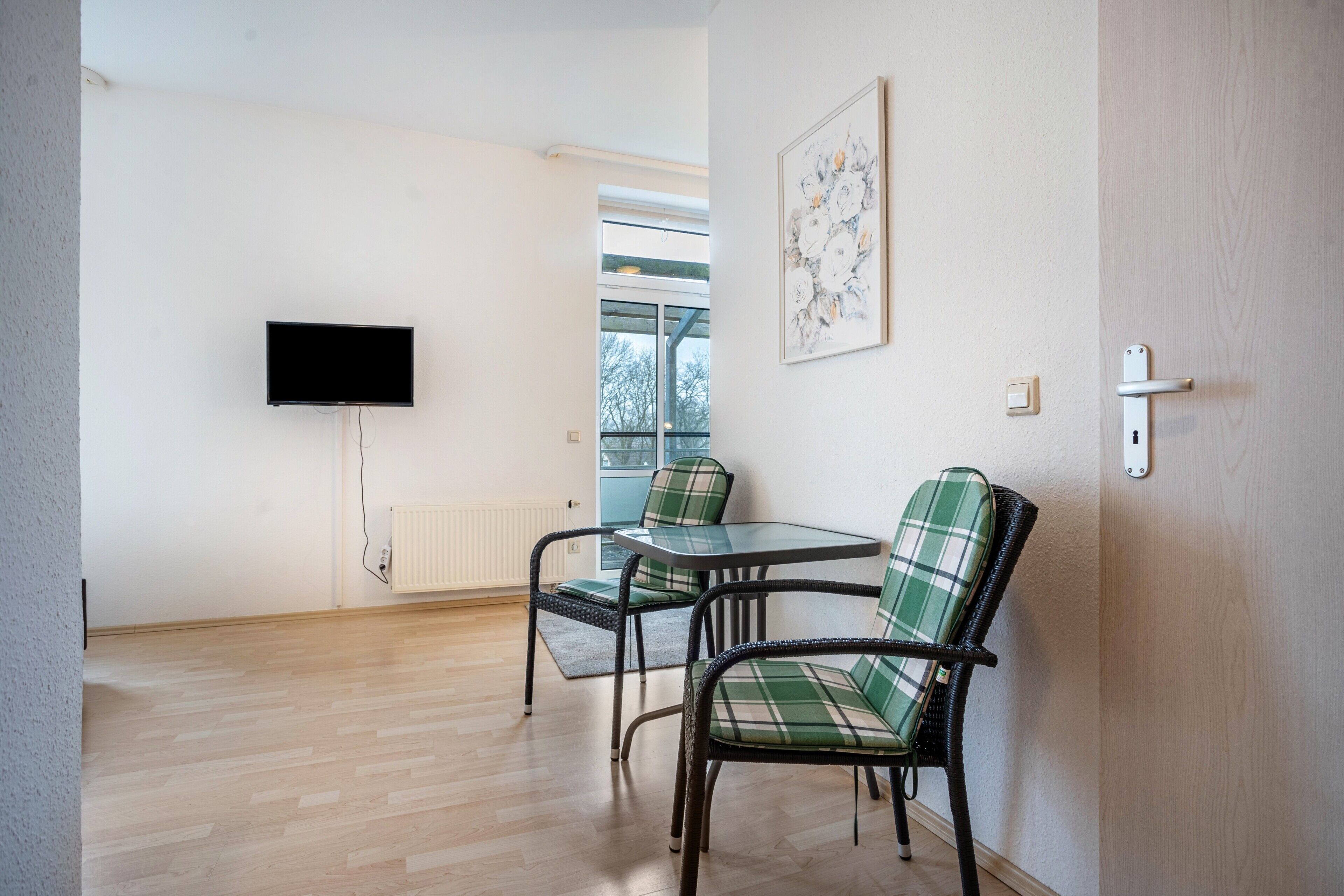 A room with a TV mounted on the wall, a window with a view, and two chairs with a table.