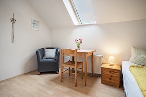 A small, well-lit room with a sloped ceiling, a desk with a lamp, a chair, a bed with a blanket, and a radiator.