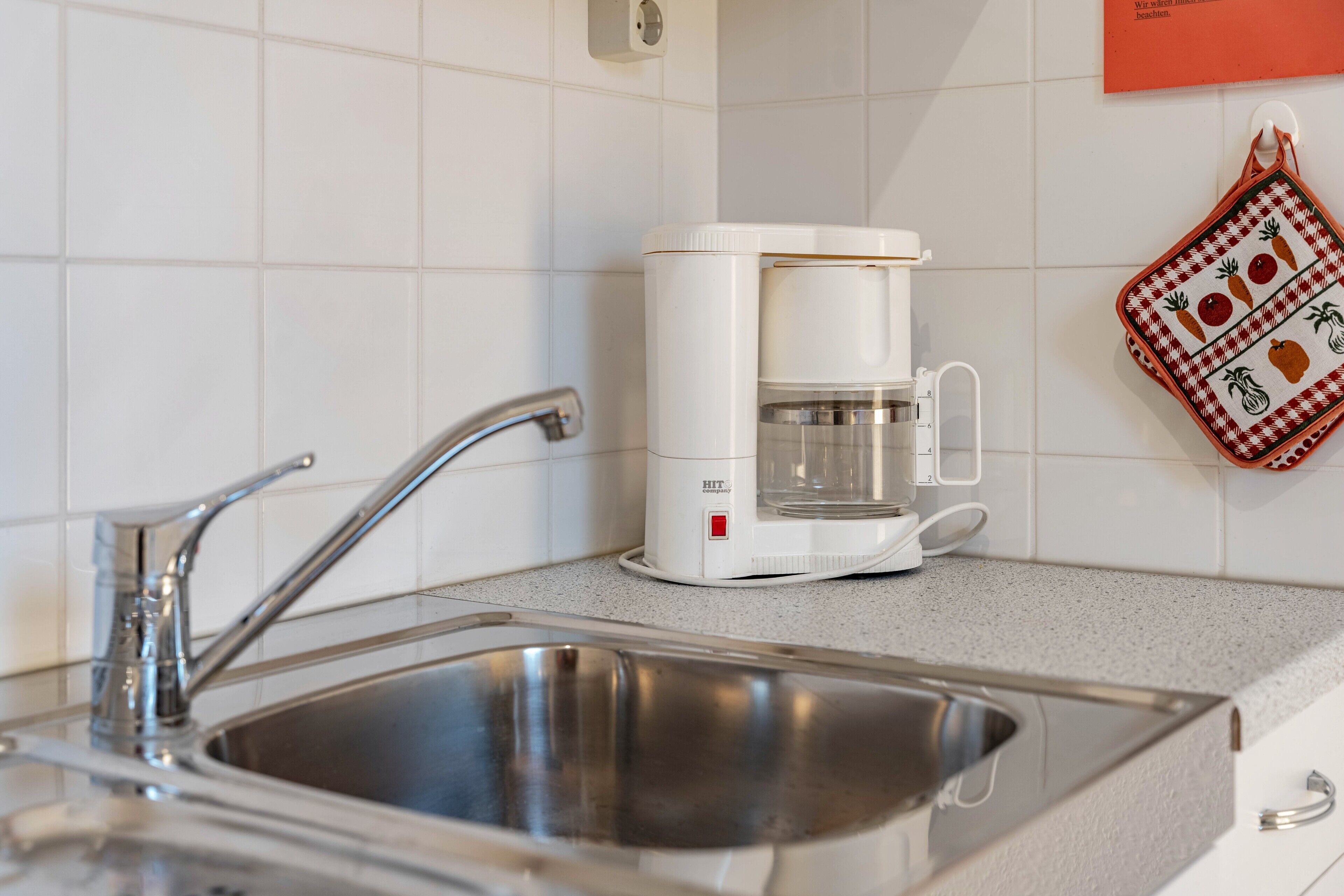 Ett kök med en diskho i rostfritt stål, en vit kaffebryggare och en röd- och vitrandig kökshandduk på väggen.