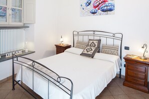 A bedroom with a metal bed frame, a radiator, a bedside table with a lamp, a wooden dresser, and a wall-mounted shelf with a book. - Ca' Du Checcu - Romantic apartment for three (Levanto)
