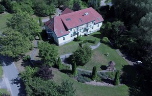 Une vue aérienne d’un grand bâtiment à deux étages, doté d’un toit rouge, entouré d’une végétation luxuriante et d’un jardin bien entretenu.