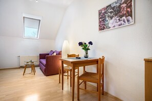 Une pièce comprenant une table à manger, des chaises, un canapé, une lampe, un vase avec des fleurs, un radiateur et une fenêtre.