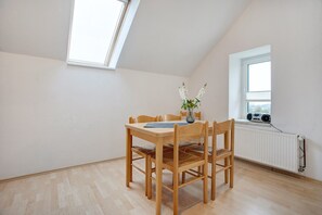 Ein Zimmer mit schrägem Dach, einem Esstisch mit Stühlen, einer Vase mit Blumen, einem Fenster mit Heizkörper und einem Radio auf der Fensterbank.