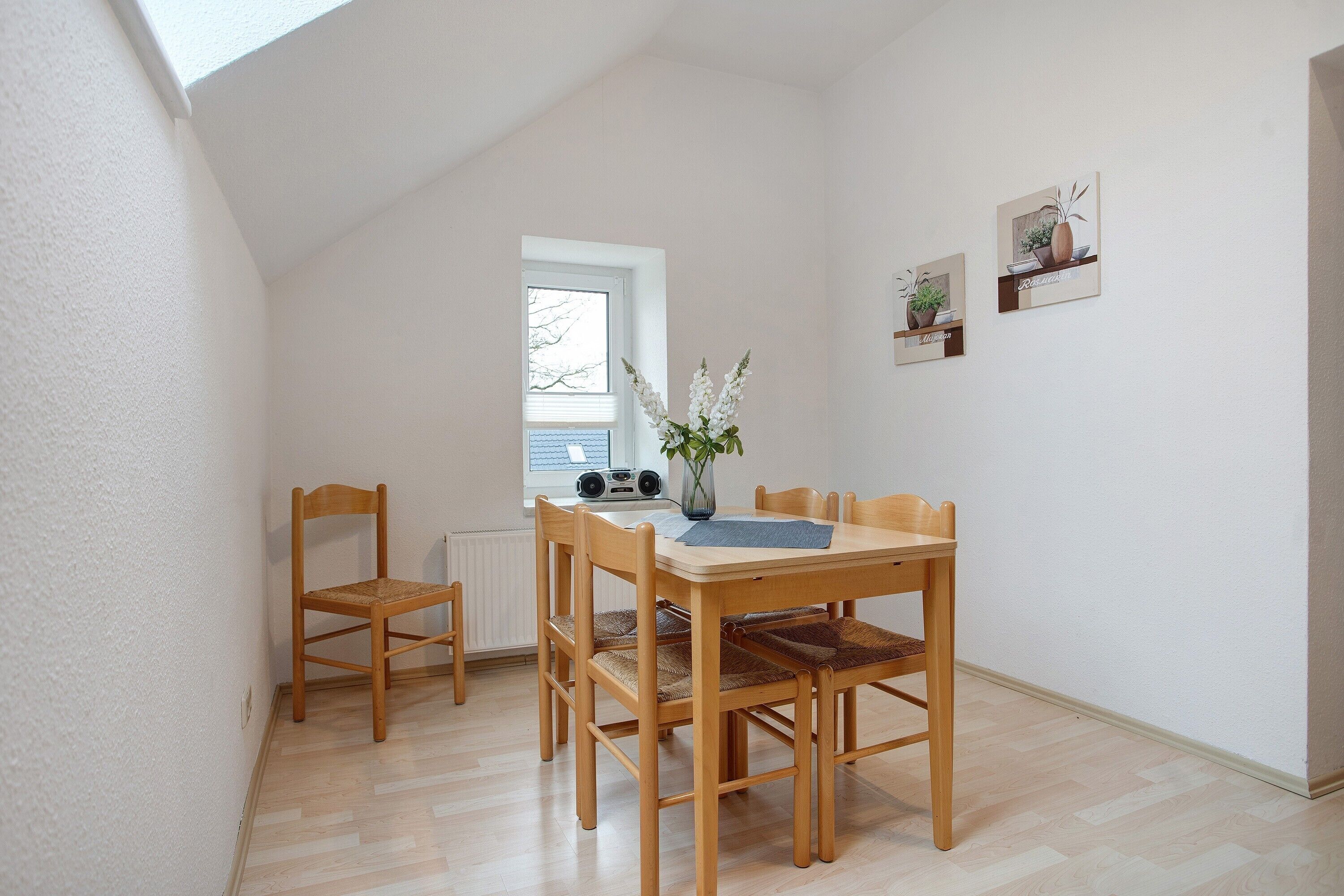 A dining area with a wooden table and chairs, a vase with flowers, a window, and a radiator.