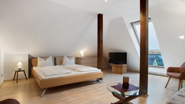 Un dormitorio moderno con cama, televisión, mesa de vidrio y vistas a una casa a través de un tragaluz.