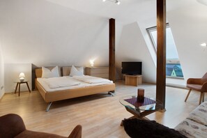Une chambre moderne avec un lit, une télévision, une table en verre et une vue sur une maison, visible à travers une lucarne.