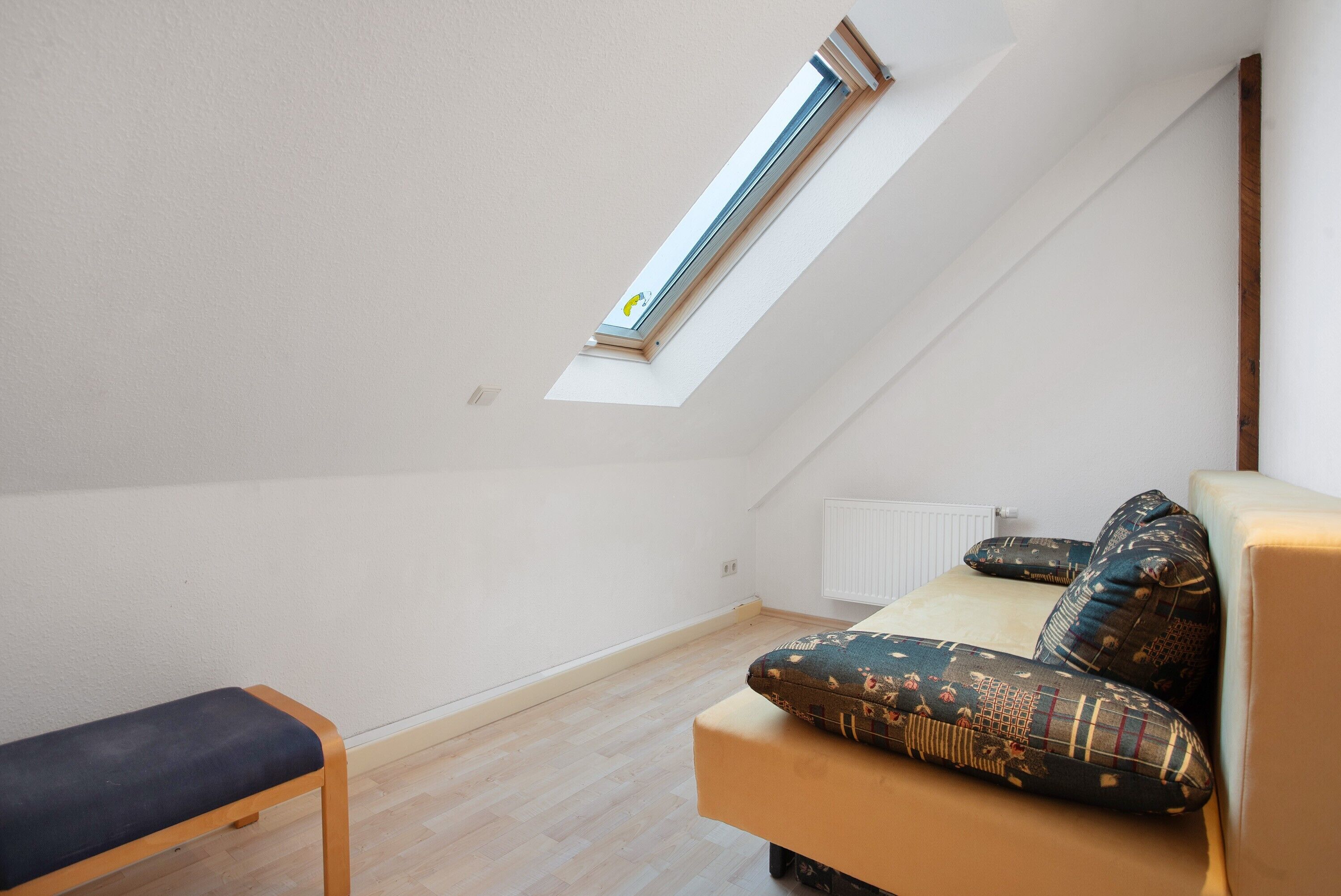 Une pièce avec un plafond en pente, un puits de lumière, un banc, un canapé avec des coussins à motifs et un radiateur.