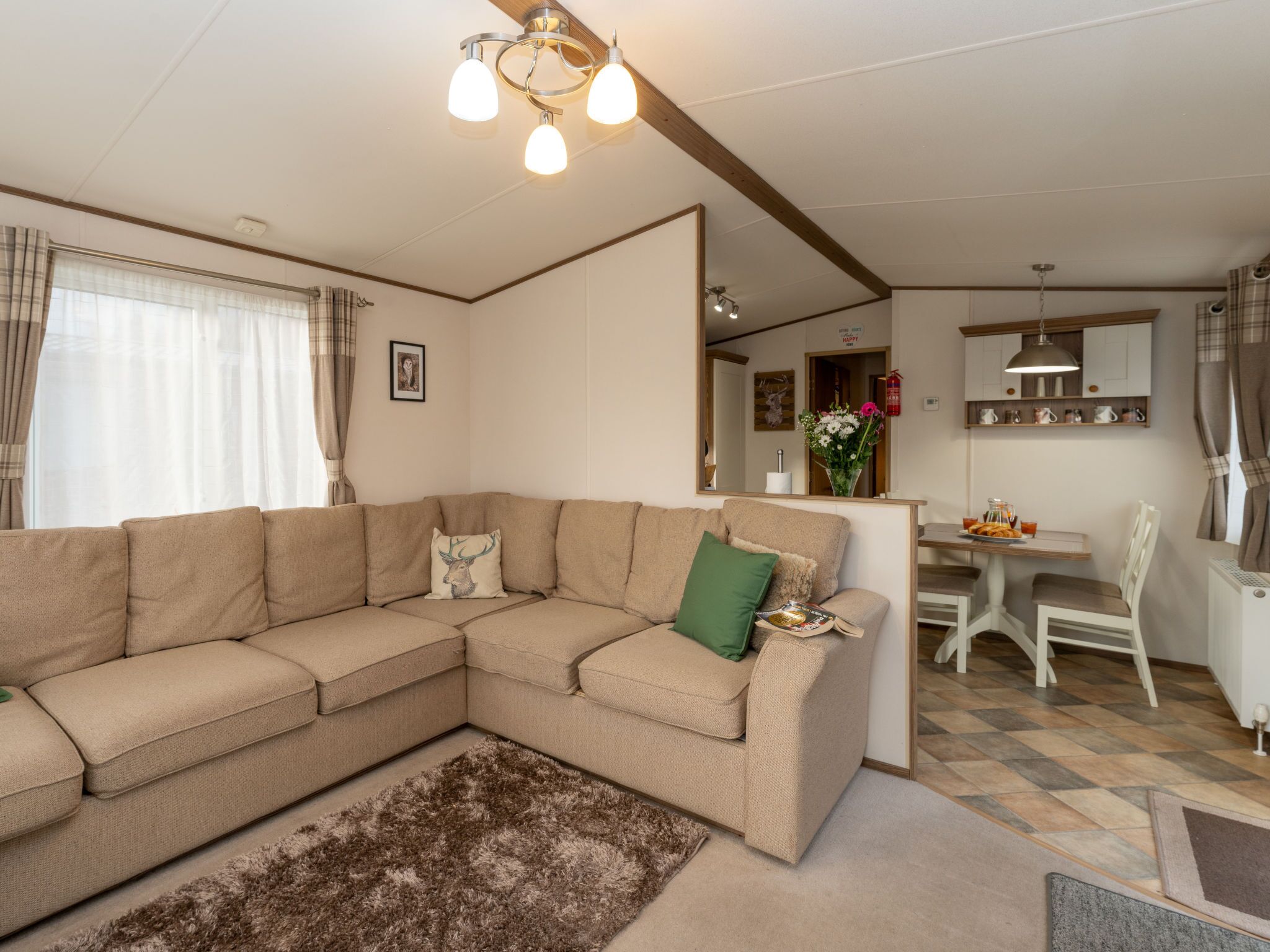 Una sala de estar con un sofá seccional color beige, un alfombra marrón mullido, una zona de comedor con mesa y sillas blancas, y una cocina con encimera y armarios.