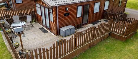A wooden cabin with a deck, a patio with chairs, and a fenced yard.