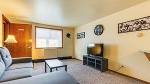 Ein Wohnzimmer mit einer Couch, einem Couchtisch, einem Fernseher auf einem Schrank, einer Uhr und einem Fenster mit Jalousien.