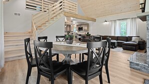 Amplia sala de estar con una escalera de madera, una mesa de comedor con sillas, una cocina al fondo y un gran ventanal con cortinas.