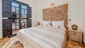 Une chambre à coucher avec un grand lit, une tête de lit décorative, une table de chevet et une fenêtre donnant sur un beau paysage.