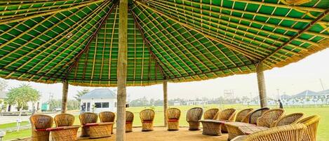An outdoor seating area with wicker furniture under a green and yellow striped roof, surrounded by a grassy field.