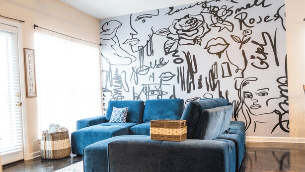 A modern living room with a blue sofa set, a decorative wall with abstract art, and a wicker basket on the floor.