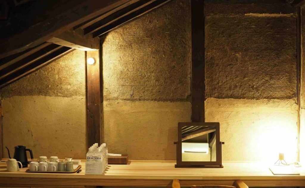 Una habitación rústica en el ático con vigas de madera a la vista, techo inclinado y un pequeño espejo sobre una estantería de madera.