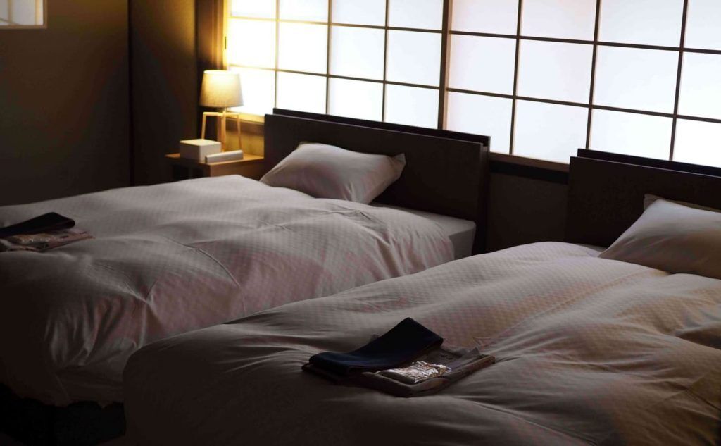 A hotel room with two beds, a nightstand with a lamp, and a window with a grid pattern.