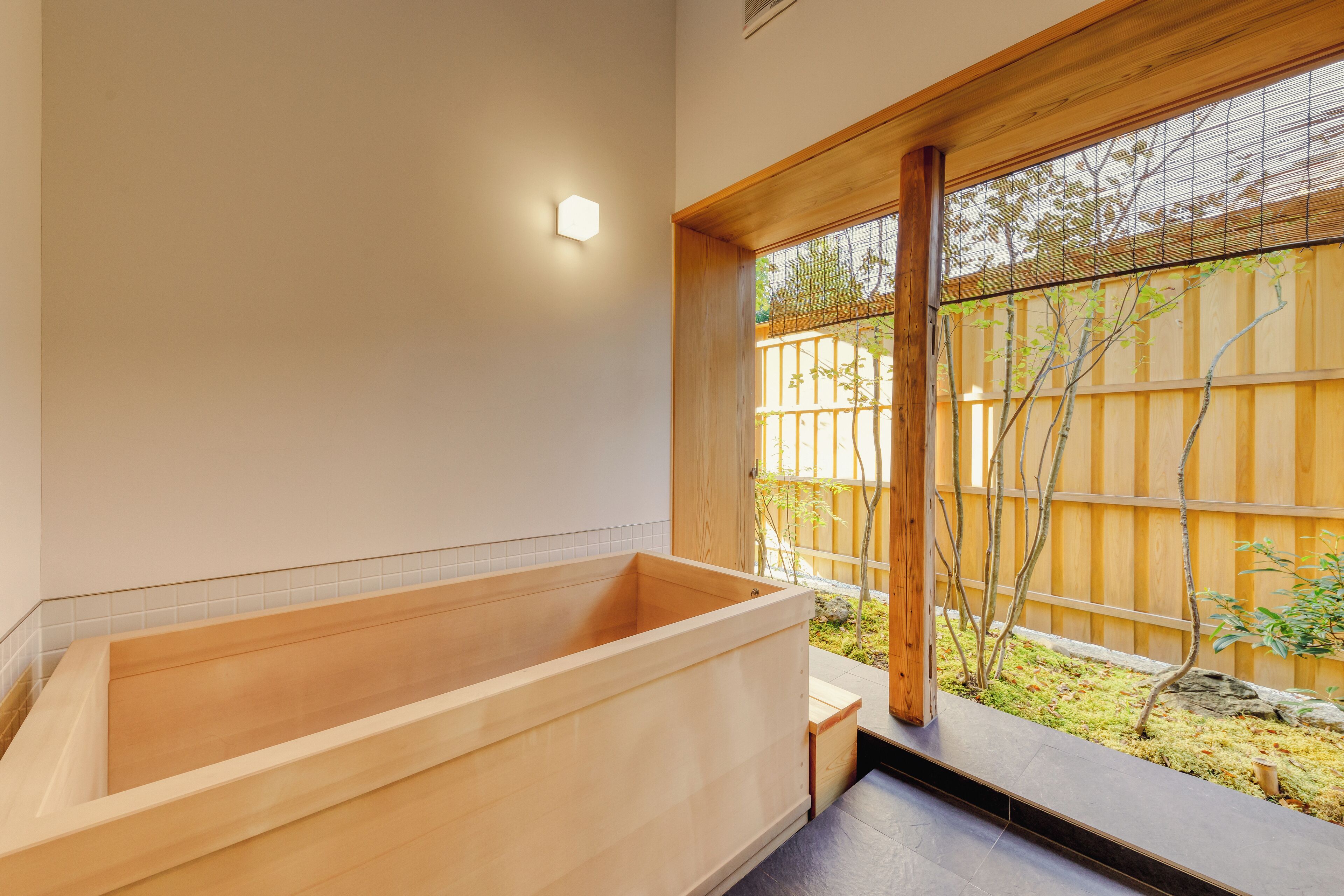 A modern bathroom with a wooden bathtub, a small window, and a view of a garden.
