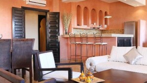 A cozy living area with a sofa, wooden chairs, and a coffee table. There is a kitchen area with a bar and chairs. - Residence Dar Lamia Marrakech: 2 Bedroom Apartment (Dar Caïd Layadi)