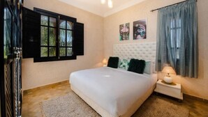 A bedroom with a white bed, a tufted headboard, a nightstand with a lamp, and a window with black shutters. - Residence Dar Lamia Marrakech: 2 Bedroom Apartment (Dar Caïd Layadi)