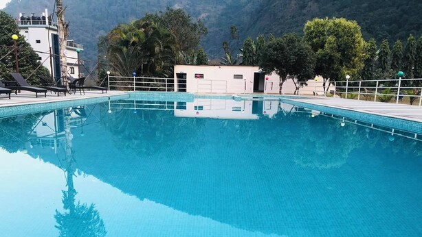 A clear blue swimming pool with a tiled deck, surrounded by trees and a mountain backdrop. - River Beach Resort (Dhussa)