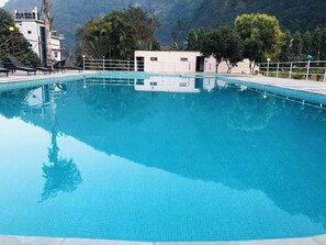 A clear blue swimming pool with a tiled deck, surrounded by trees and a mountain backdrop. - River Beach Resort (Dhussa)