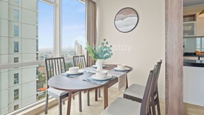 A dining area with a round table, chairs, a vase with green leaves, and a city view through large windows. - Canna by Kozystay - Menteng (Jakarta)