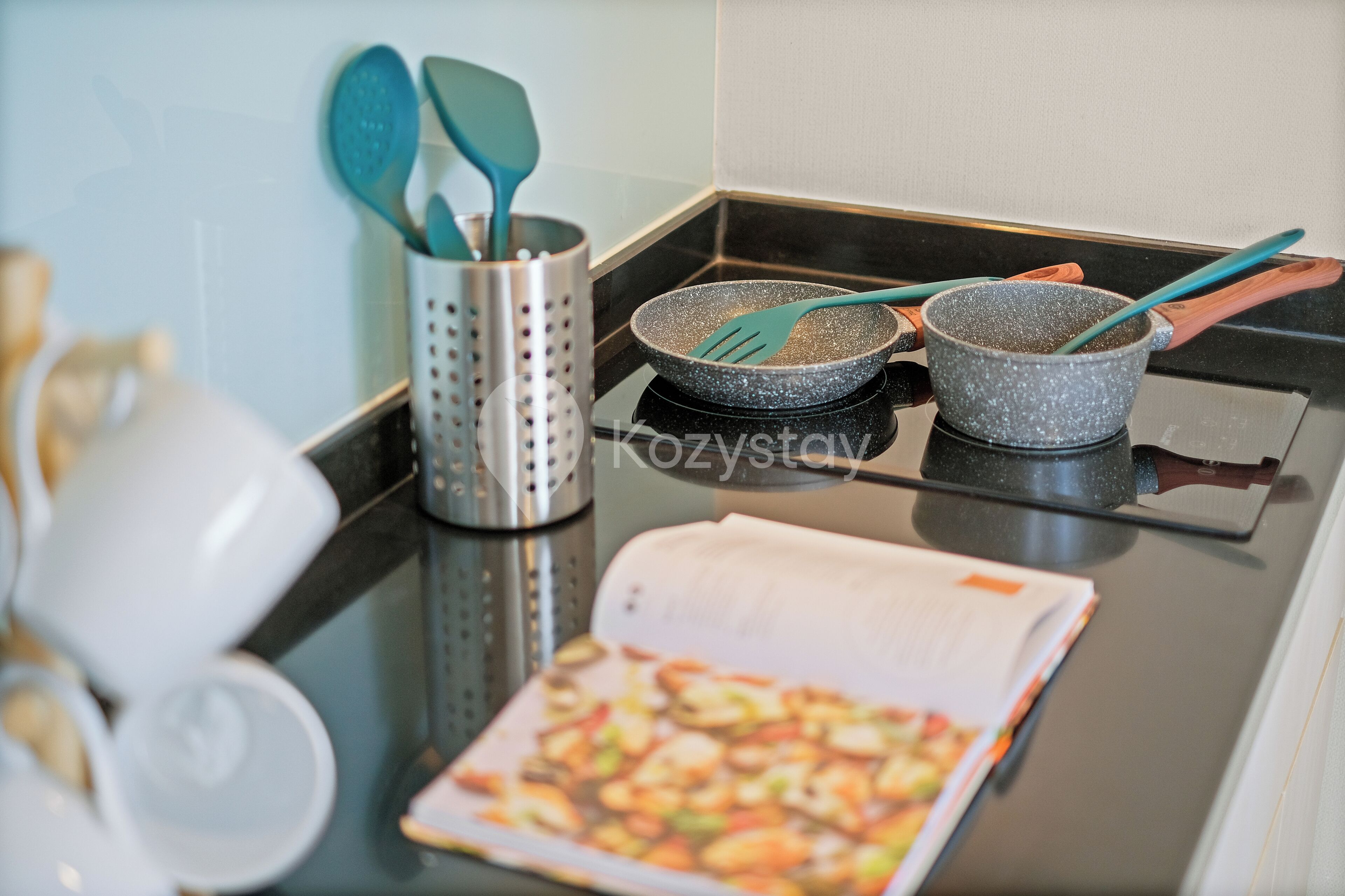 A kitchen counter with a cookbook, a frying pan, and a pot with a wooden handle.