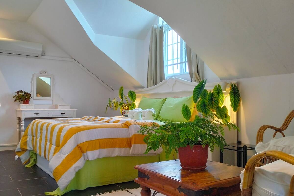 Une chambre avec un plafond en pente, un lit avec une literie à rayures, une table de chevet avec un pot de plante et une fenêtre avec des rideaux.