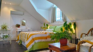 Een slaapkamer met een schuine balkenlaag, een bed met gestreepte beddengoed, een nachtkastje met een potplant en een raam met gordijnen.