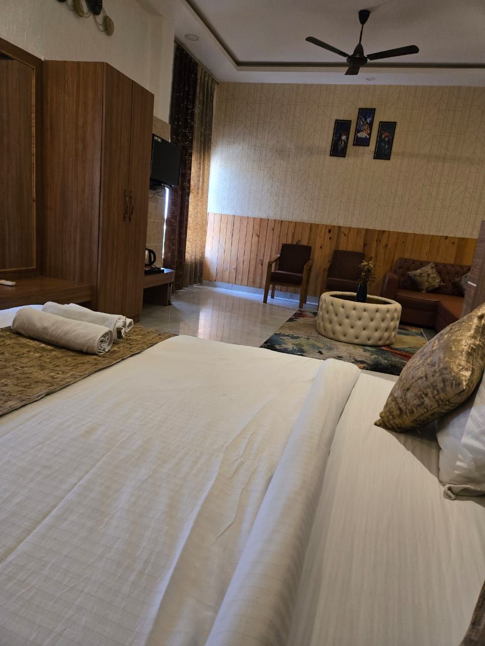 A hotel room with two beds, a wooden wardrobe, a ceiling fan, and a seating area with a sofa and armchairs.