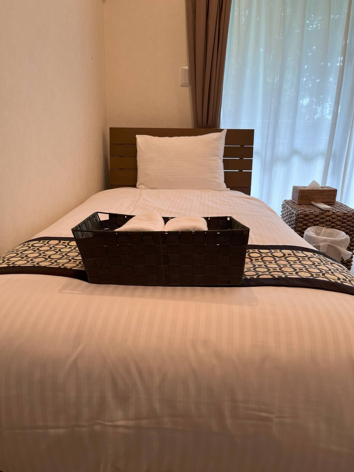 A neatly made bed with a white pillow and a brown headboard. A black woven basket is placed on the bed.